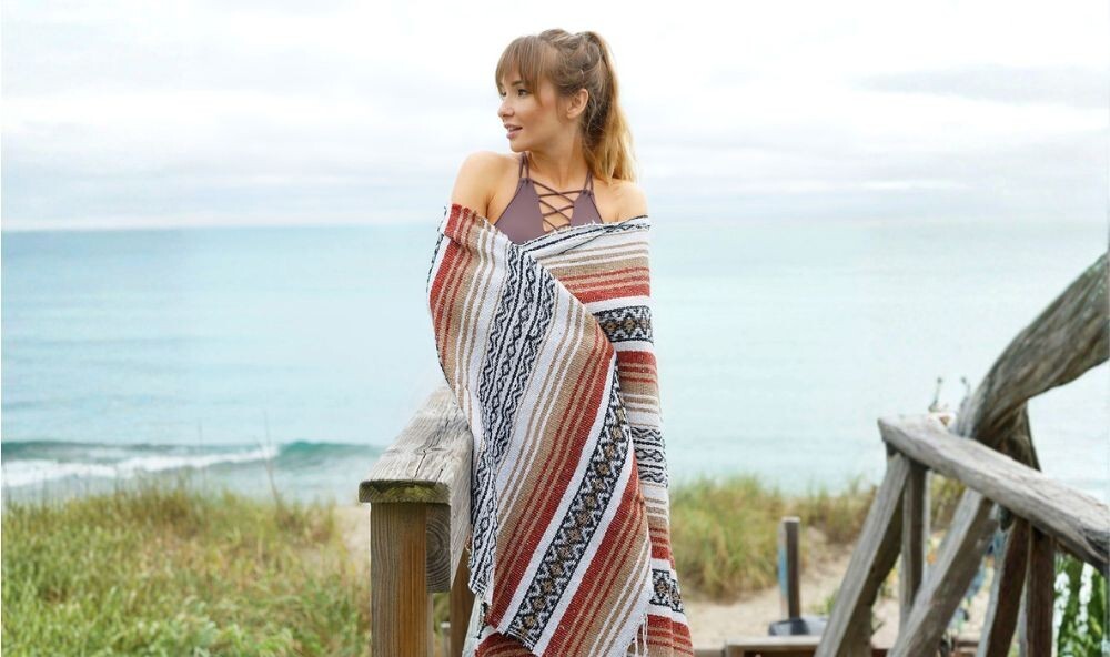 Woman wrapped in a Mexican blanket by the beach, adding warmth over leggings