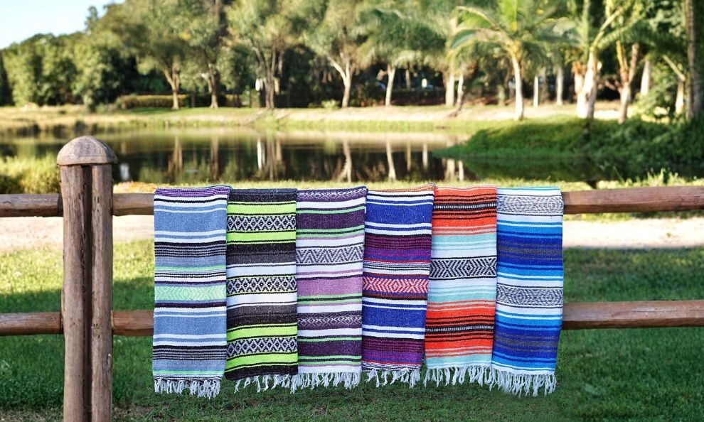 A row of colorful woven recycled blankets hanging on a wooden fence by a lakeside