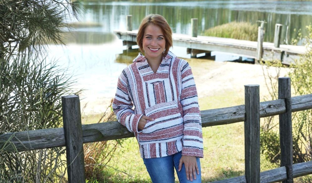 Woman wearing a recycled-fiber Baja hoodie outdoors by a lakeside wooden fence