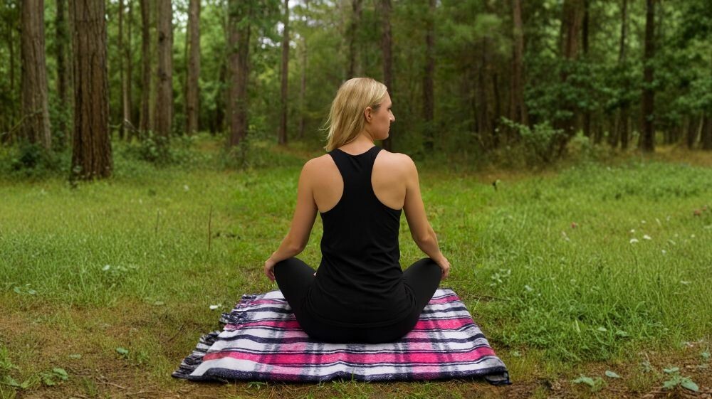 Woman meditating on a Svaroopa yoga blanket in a forest clearing, seated cross-legged on a pink and black woven mat.