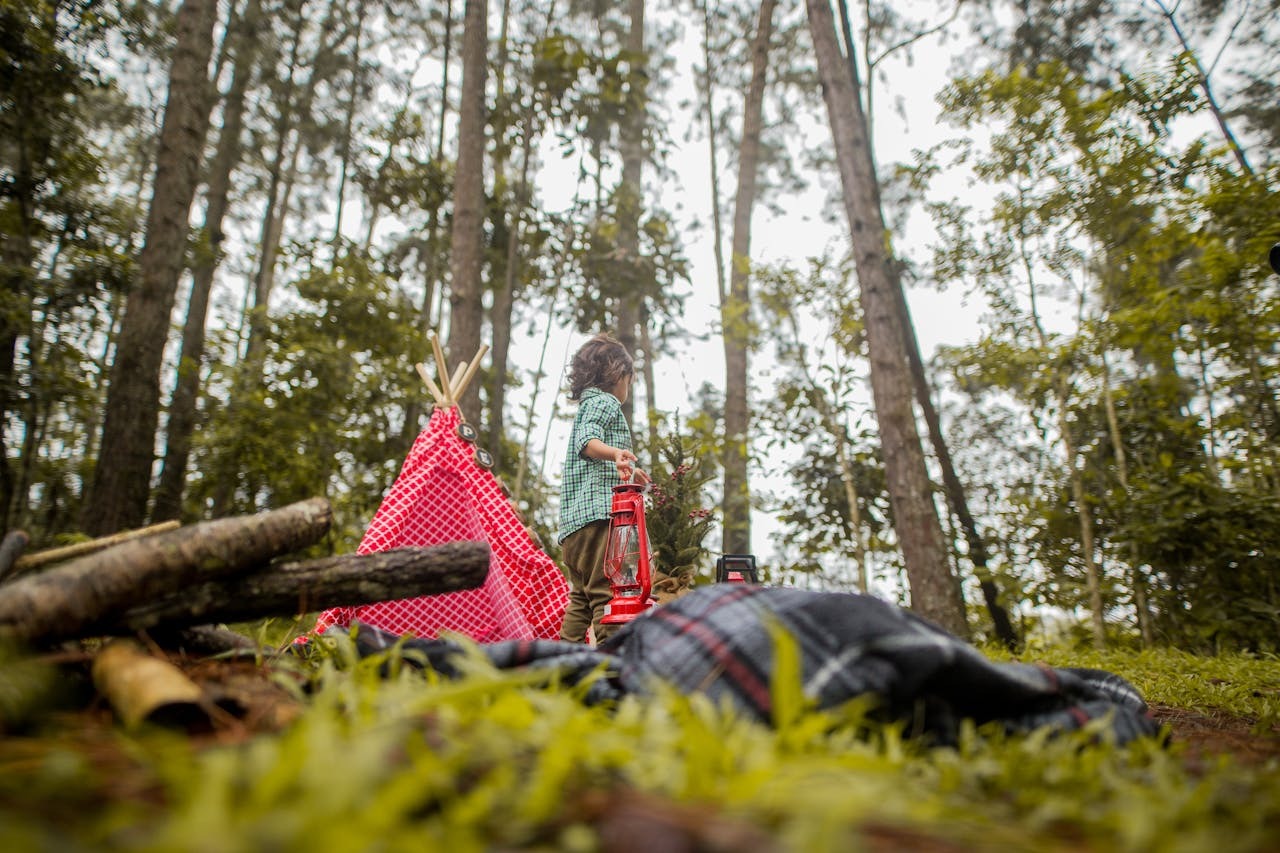 Child camping in a forest with blankets, firewood, and a lantern, illustrating eco-friendly outdoor gear